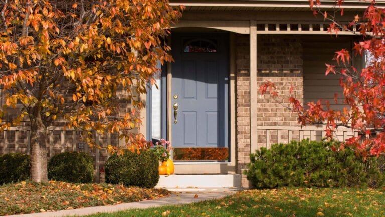 A house entrance decorated with pumpkins and surrounded by vibrant autumn foliage.