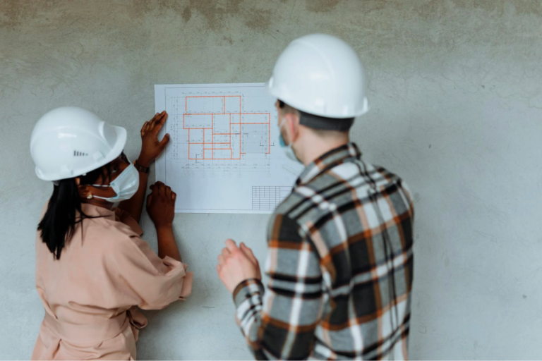 Two people in hard hats are holding up a blueprint against a wall.